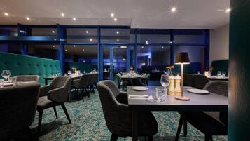 Een restaurant met een modern interieur, met een smaragdgroene gestoffeerde zithoek, donkere stoelen en tafels gedekt met borden en glazen.