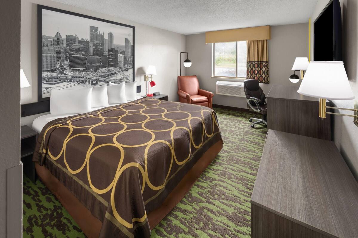 A hotel room with a large bed, a desk, a chair, a desk lamp, and a cityscape painting on the wall.
