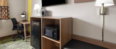 A hotel room with a desk, a microwave, a television, a chair, and a bench.