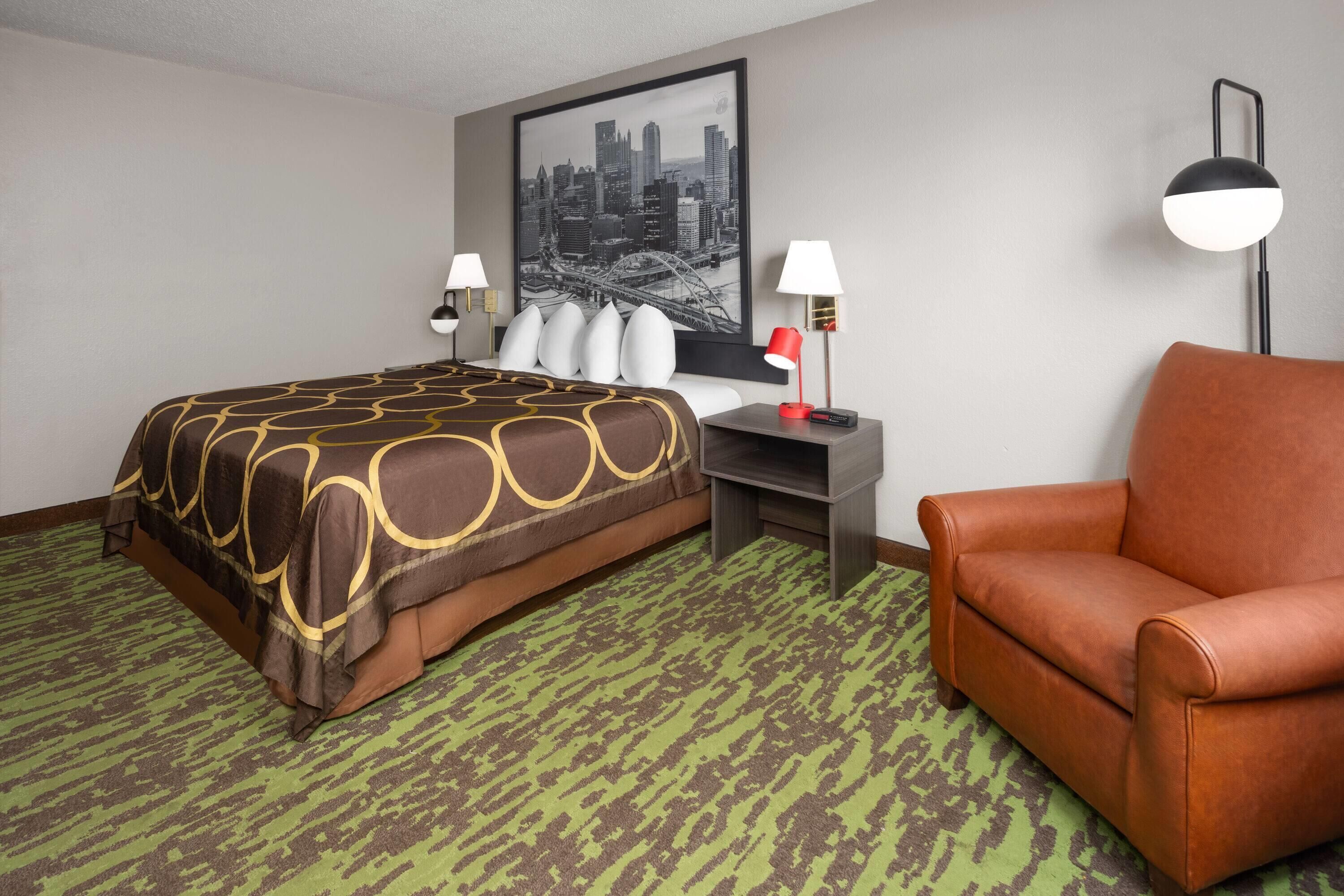 A hotel room with a bed, a brown leather armchair, a desk with a lamp, and a cityscape picture on the wall.