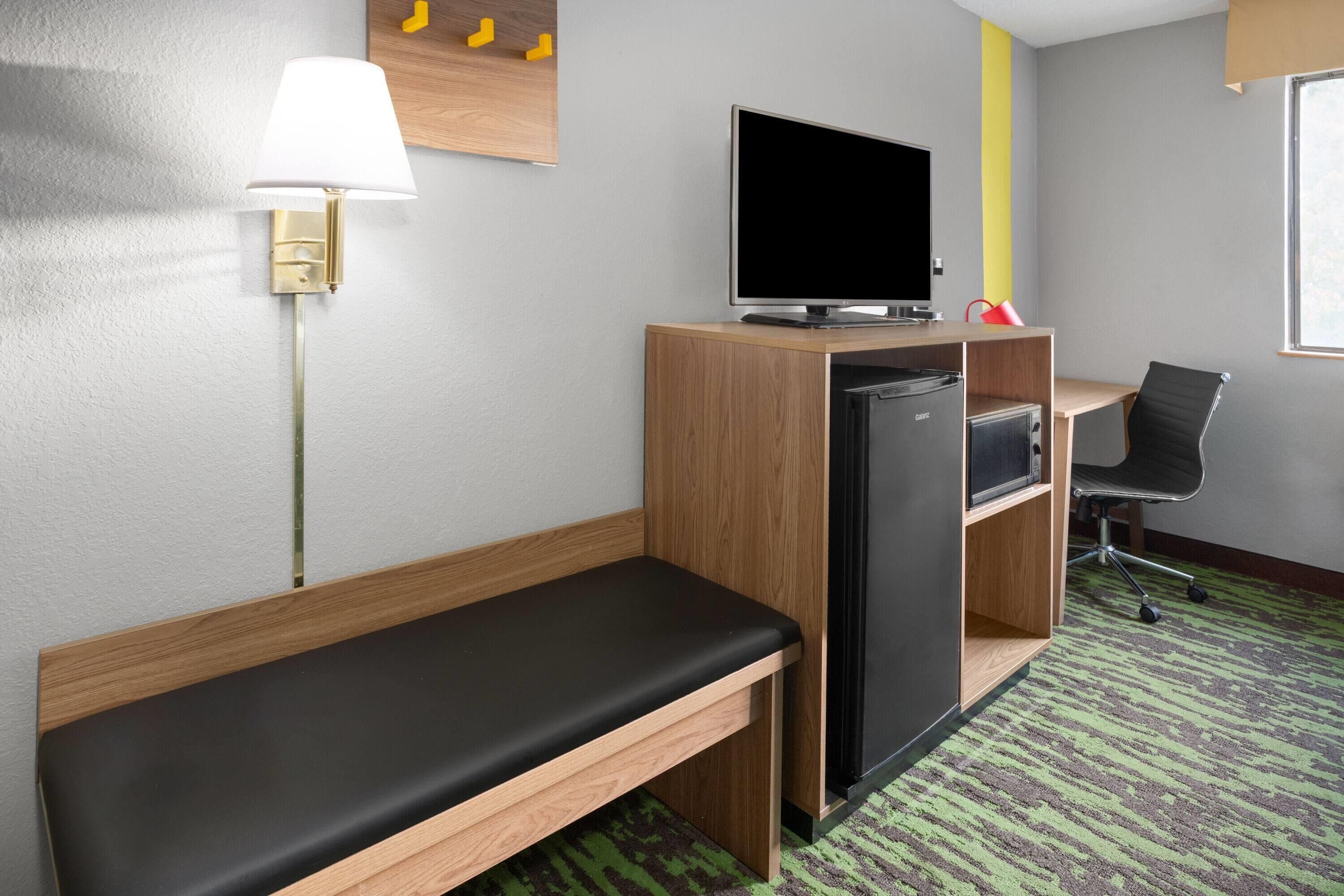 Habitación de hotel compacta con una cama individual, un escritorio con ordenador, televisor y un pequeño refrigerador.