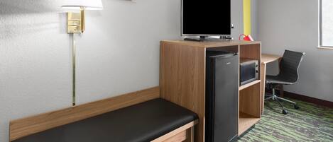 A compact hotel room with a single bed, a desk with a computer, a television, and a small refrigerator.