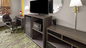 Habitación de hotel con televisor de pantalla plana, escritorio con lámpara, silla, microondas y cafetera.