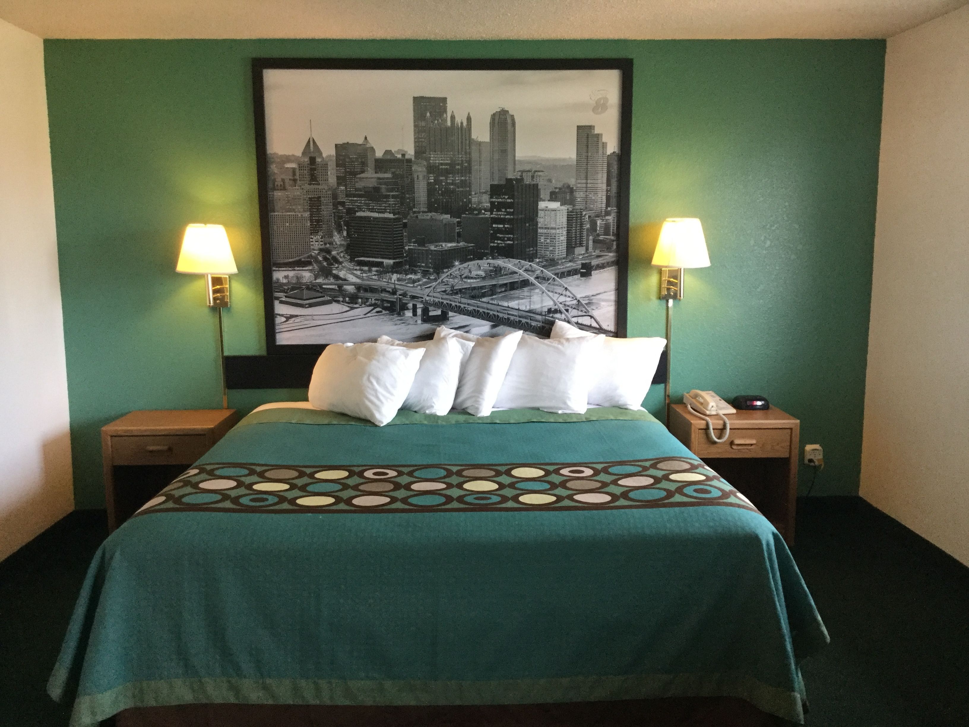 A hotel room with a bed, two bedside lamps, a framed cityscape picture on the wall, and a telephone on the nightstand.