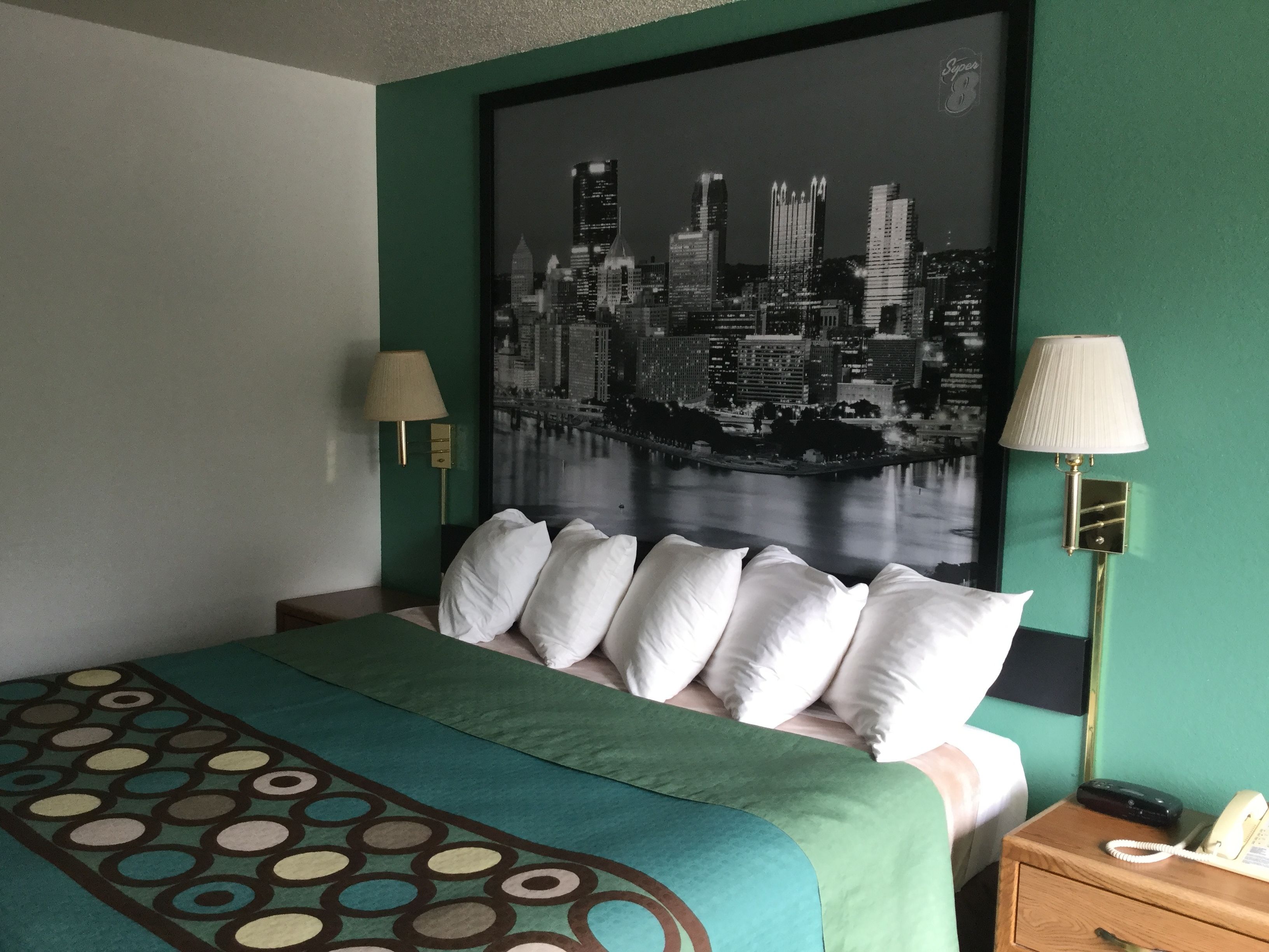 Hotelkamer met een bed, groene wanden, een stadsgezicht schilderij, twee bedlampen en een telefoon op een houten nachtkastje.