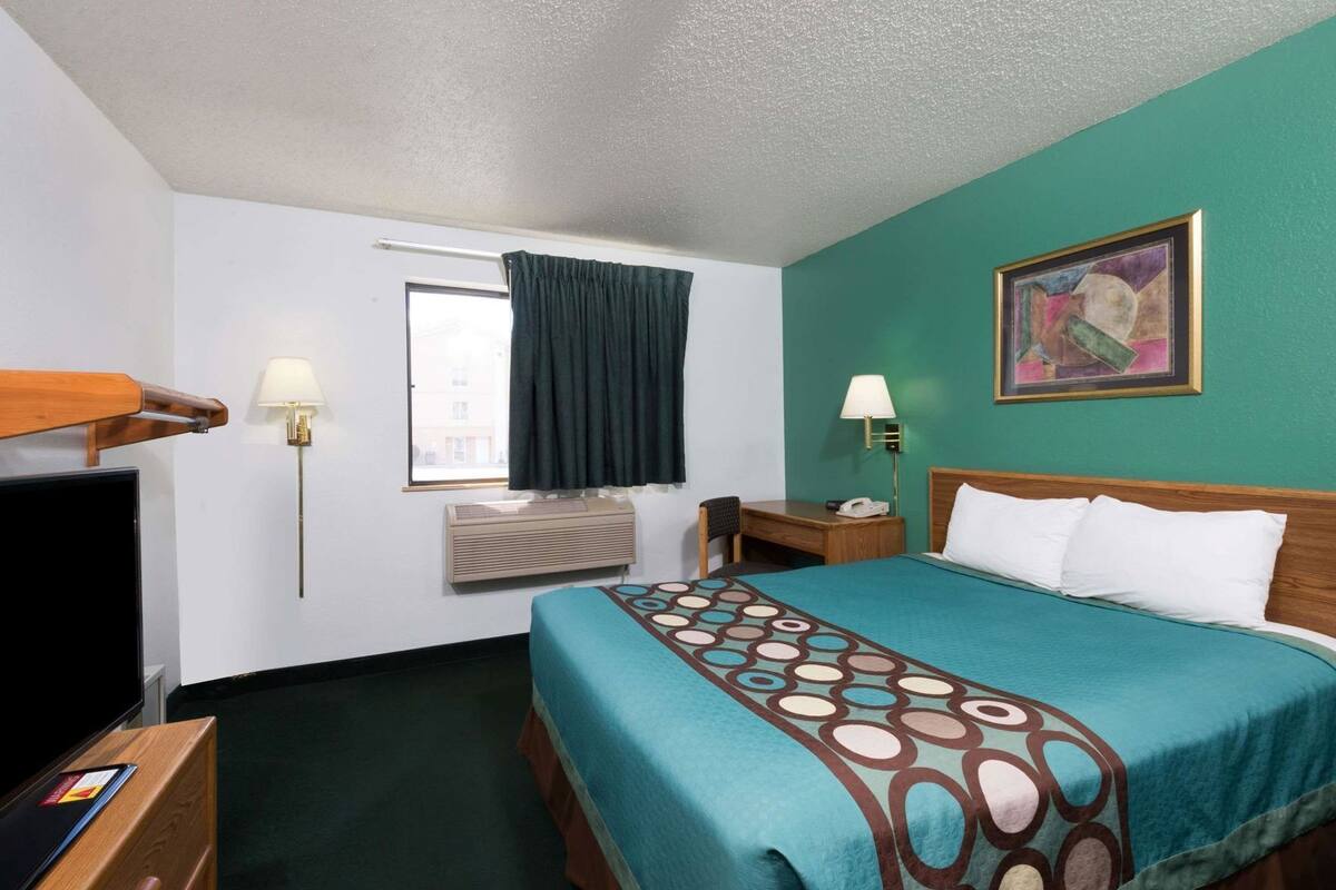 A hotel room with a bed, a desk, a chair, a TV, a window with curtains, an air conditioner, and a painting on the wall.