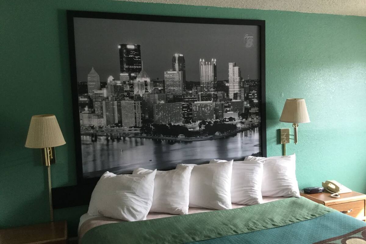 A hotel room with a bed, two bedside lamps, a framed cityscape picture, and a green wall.