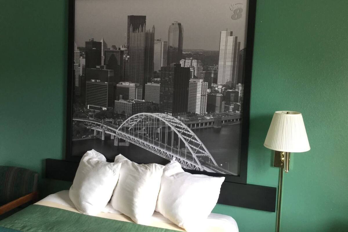 A framed cityscape photograph with a bridge, a bed with pillows, and a lamp on a green wall.