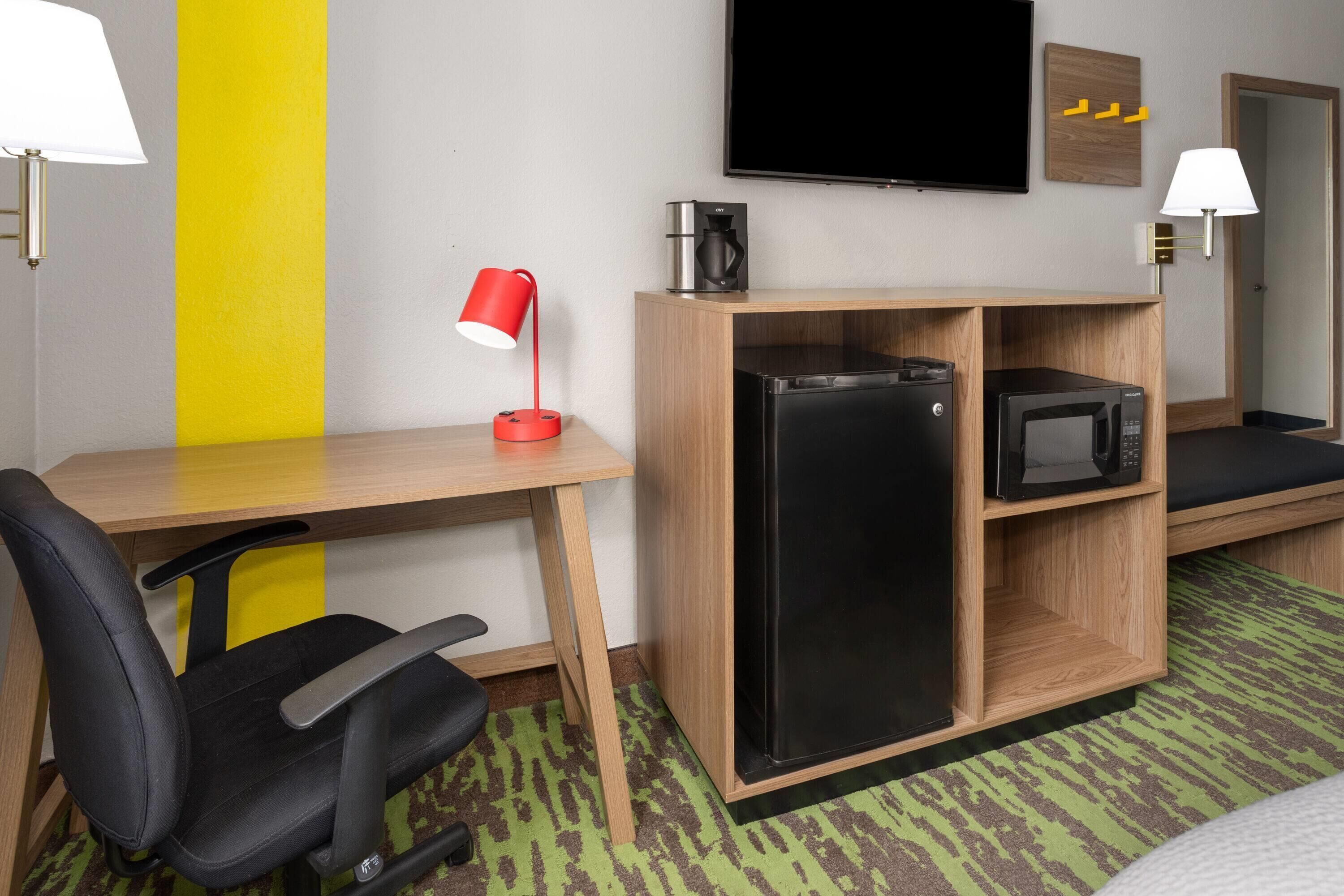 Habitación de hotel con escritorio de madera, silla de oficina negra, lámpara de escritorio roja, refrigerador negro, microondas, televisor de pantalla plana montado en la pared y alfombra verde con estampado.