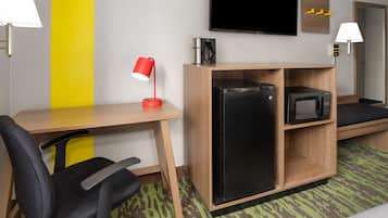 Quarto de hotel com escrivaninha de madeira, cadeira de escritório preta, luminária de mesa vermelha, geladeira preta, micro-ondas, TV de tela plana na parede e tapete verde estampado.