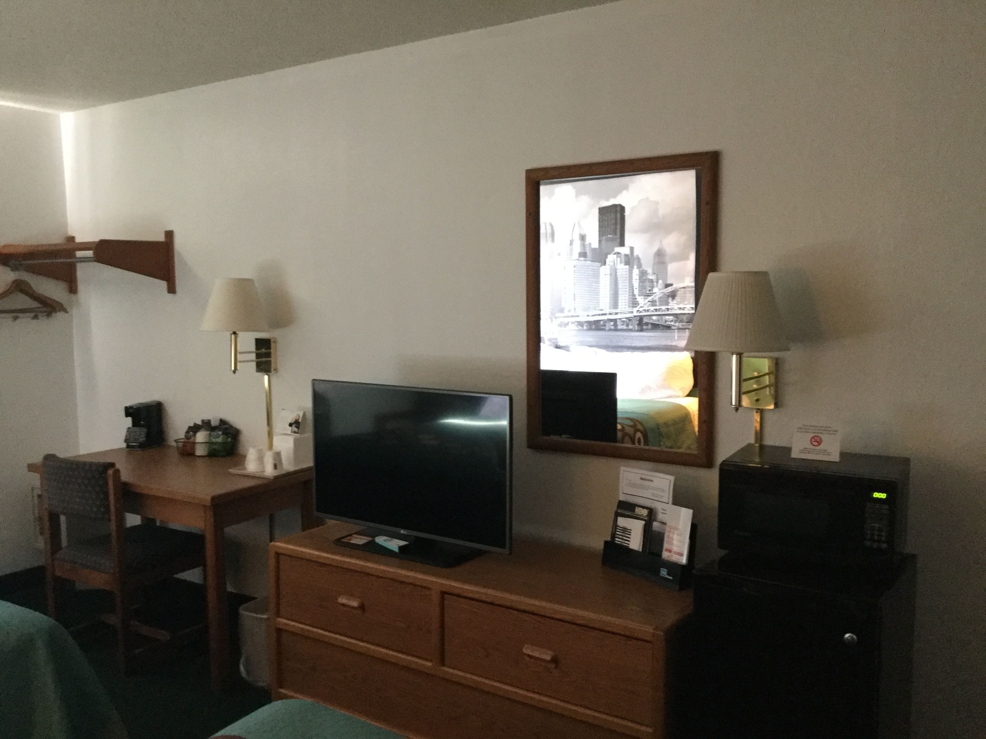 A hotel room with a TV, desk, chair, and a mirror reflecting a cityscape.