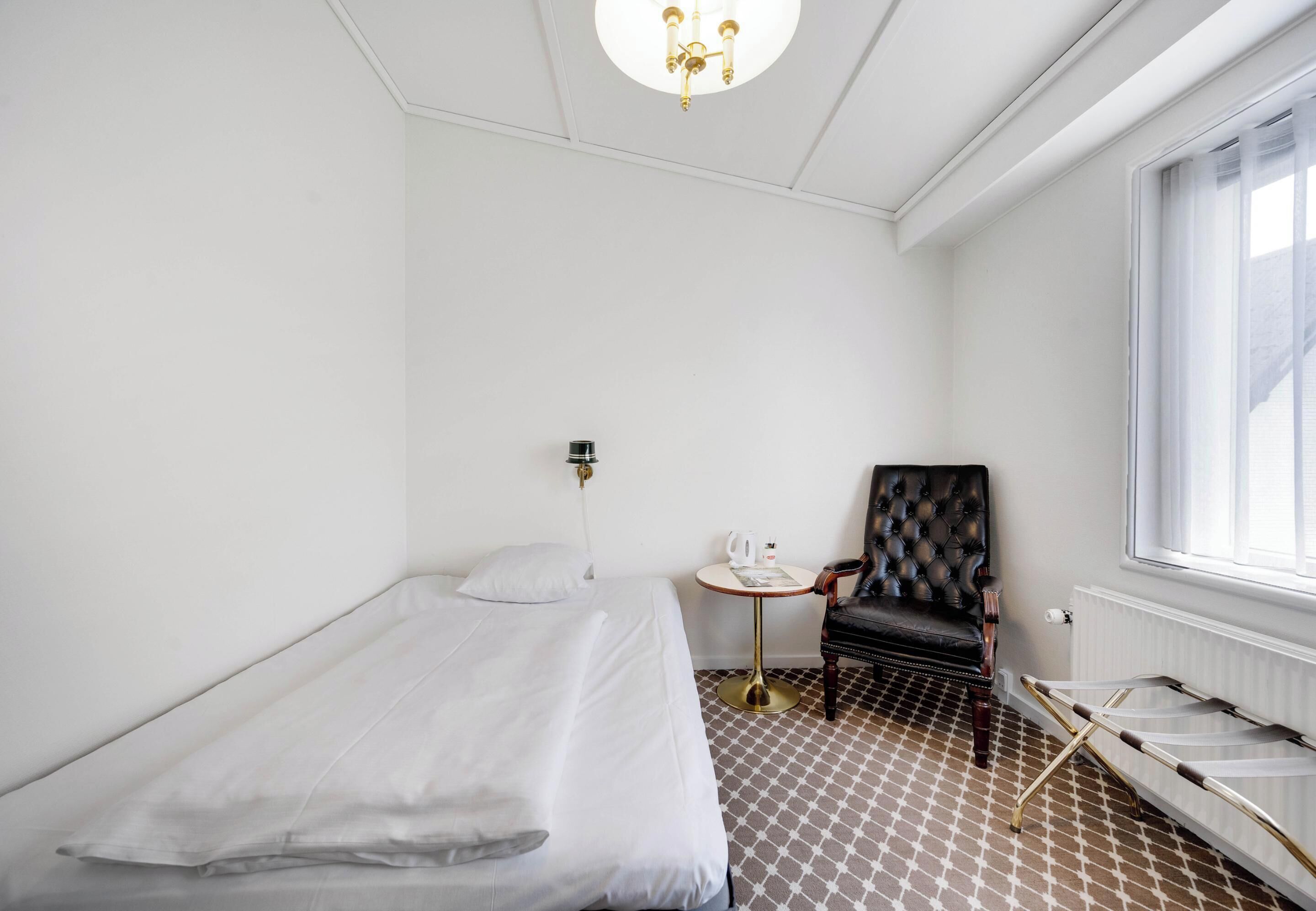 Ein Hotelzimmer mit einem Einzelbett, einem kleinen runden Tisch mit Wasserkocher, einem Sessel und einer Heizung.