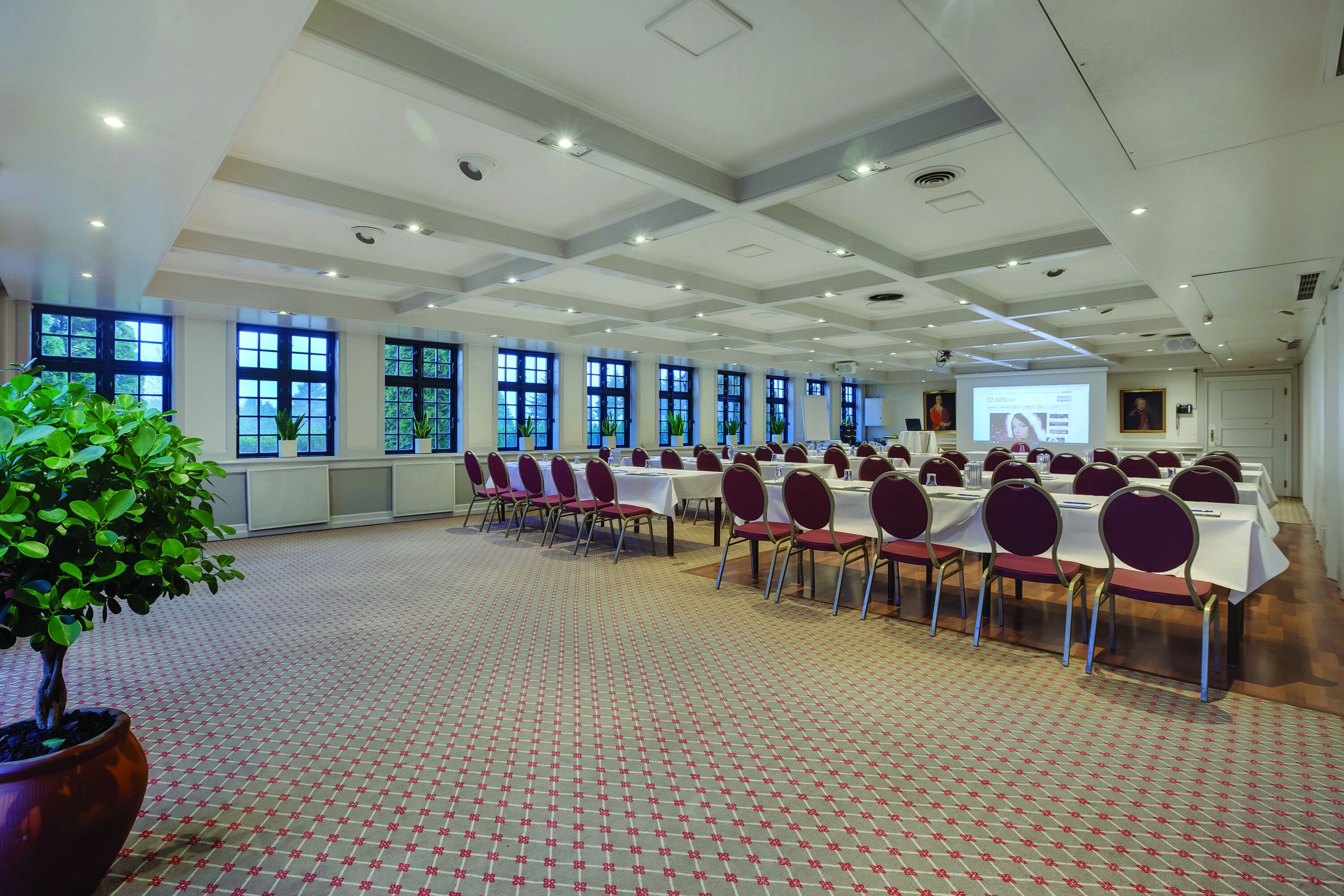 Un salón de conferencias con mesas, sillas, ventanas amplias y una pantalla de proyector.