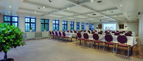 Un salón de conferencias con mesas, sillas, ventanas amplias y una pantalla de proyector.