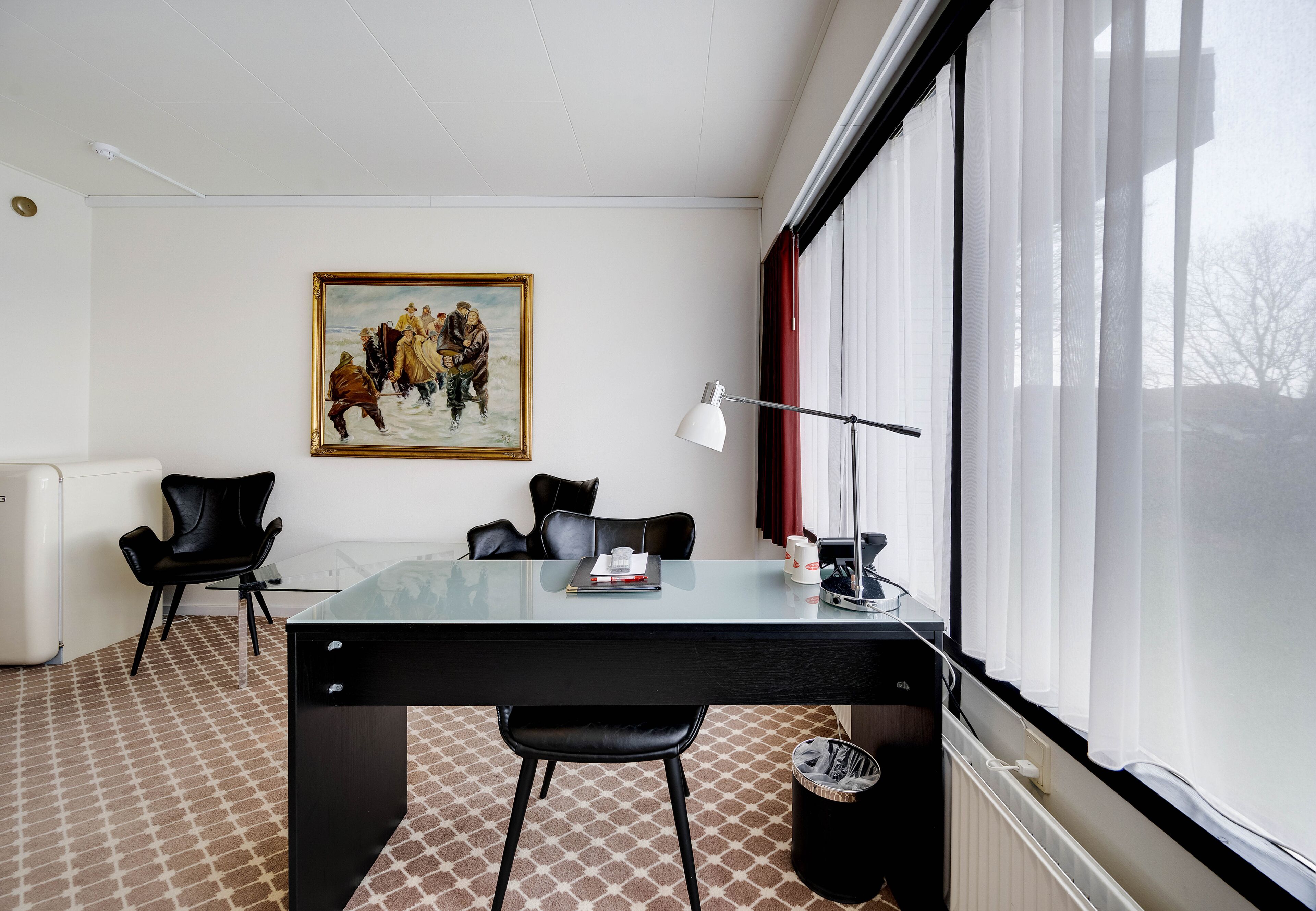 A modern hotel room with a glass table, black chairs, a painting on the wall, and a large window with curtains.