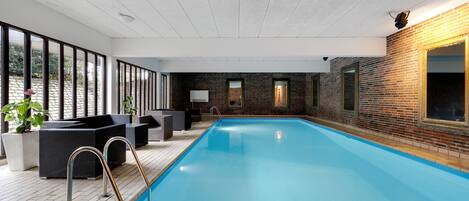 Una piscina interior con cubierta de madera, zona de estar y grandes ventanales.