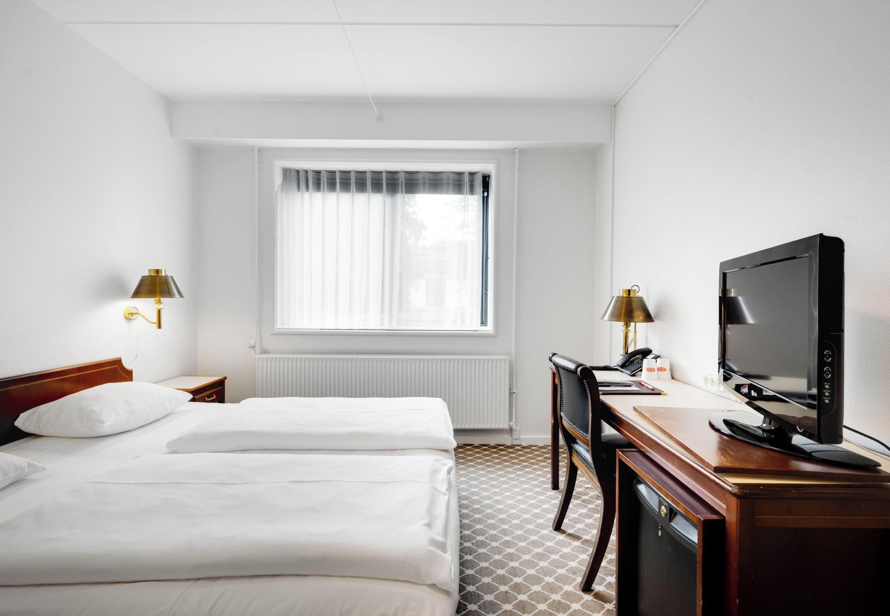 Et hotelværelse med to senge, et skrivebord med computer, et fjernsyn og et vindue med gardiner.