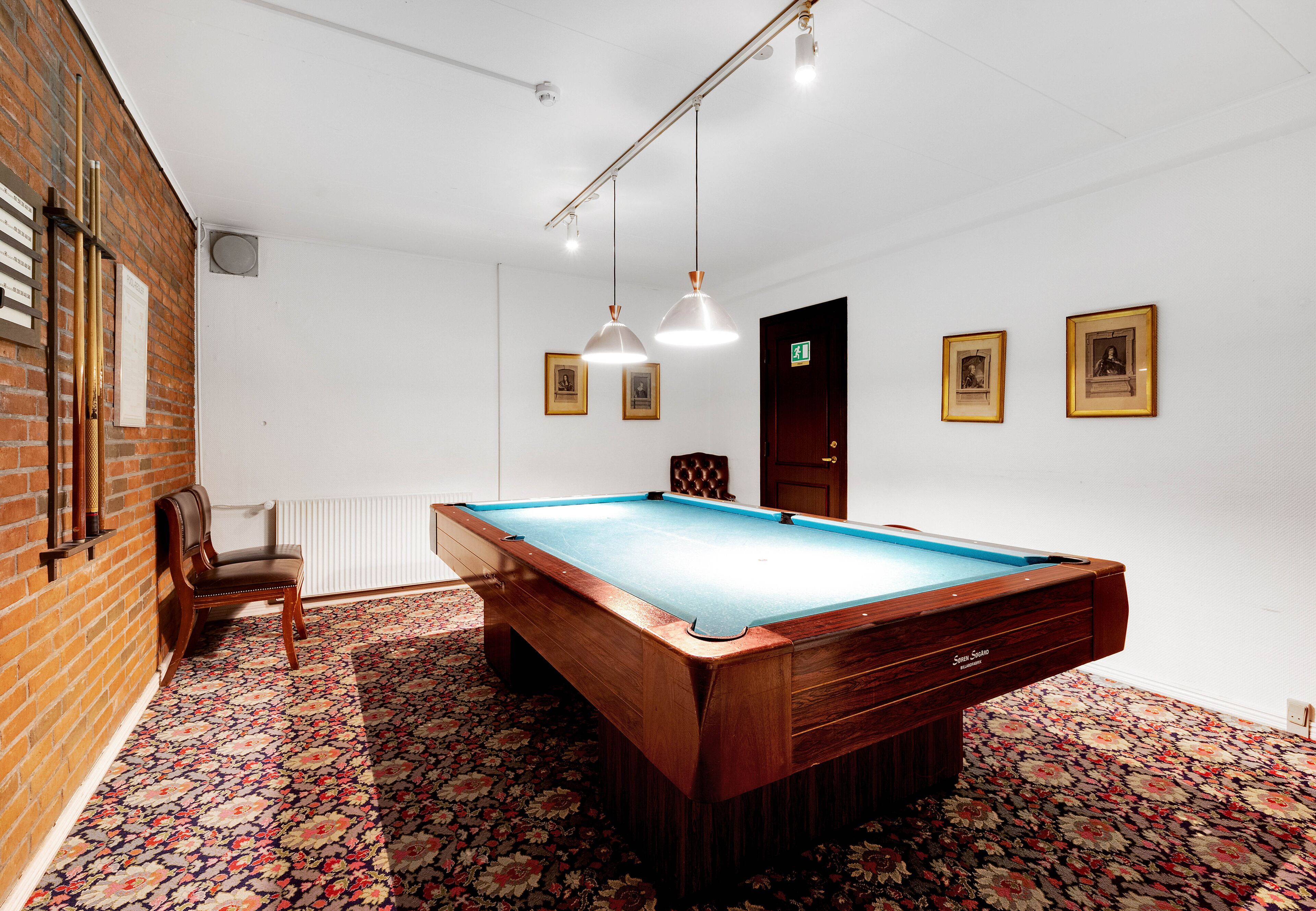 A pool table in a room with a floral carpet, a brick wall, and framed pictures on the wall.