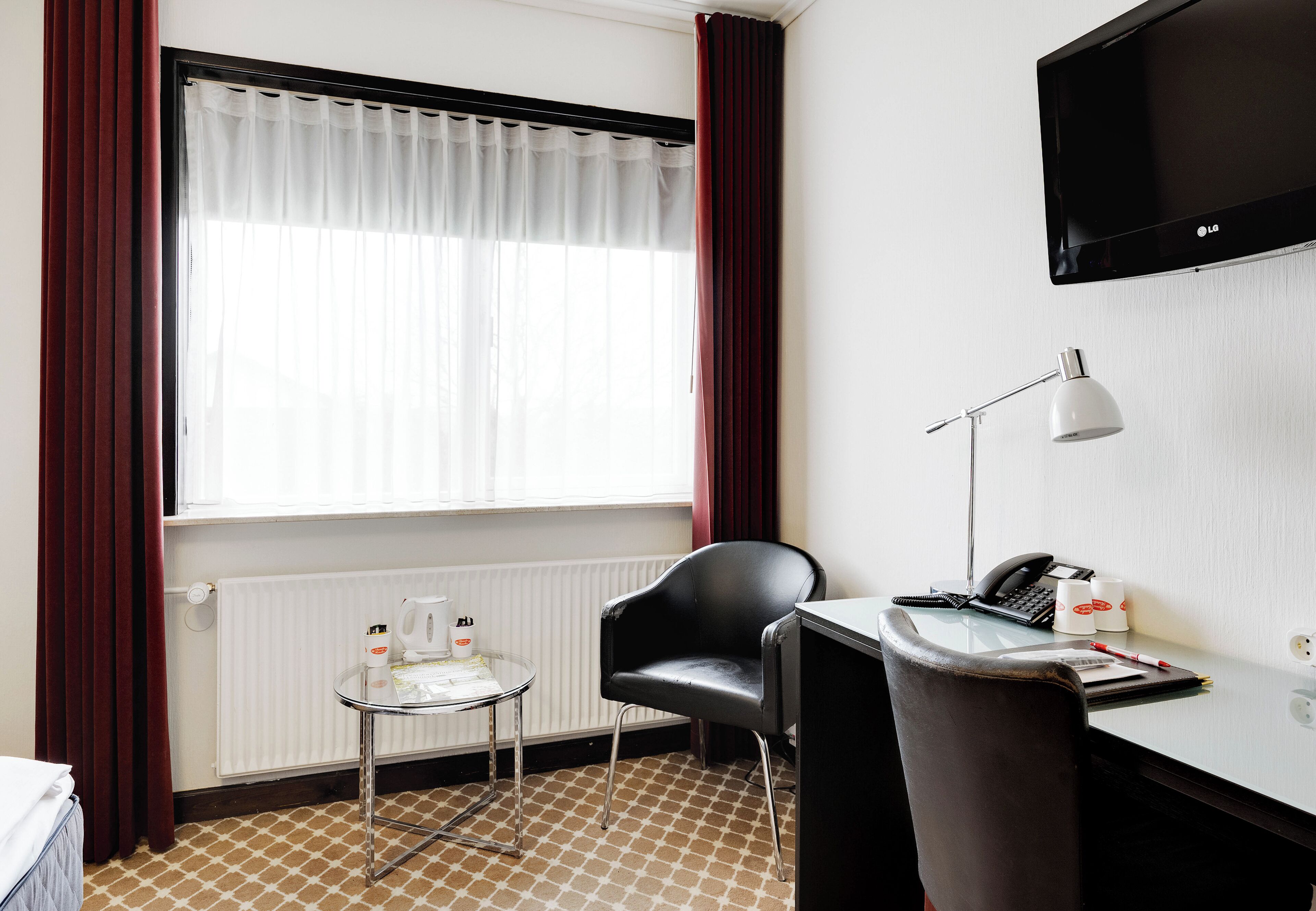 A hotel room with a bed, a desk with a chair, a small table with a lamp, a TV, and a window with curtains.