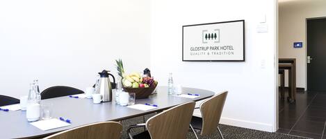 A conference room with a long table, four chairs, a fruit bowl, water bottles, and a coffee pot. A framed sign on the wall reads "GLOSTRUP PARK HOTEL QUALITY & TRADITION".