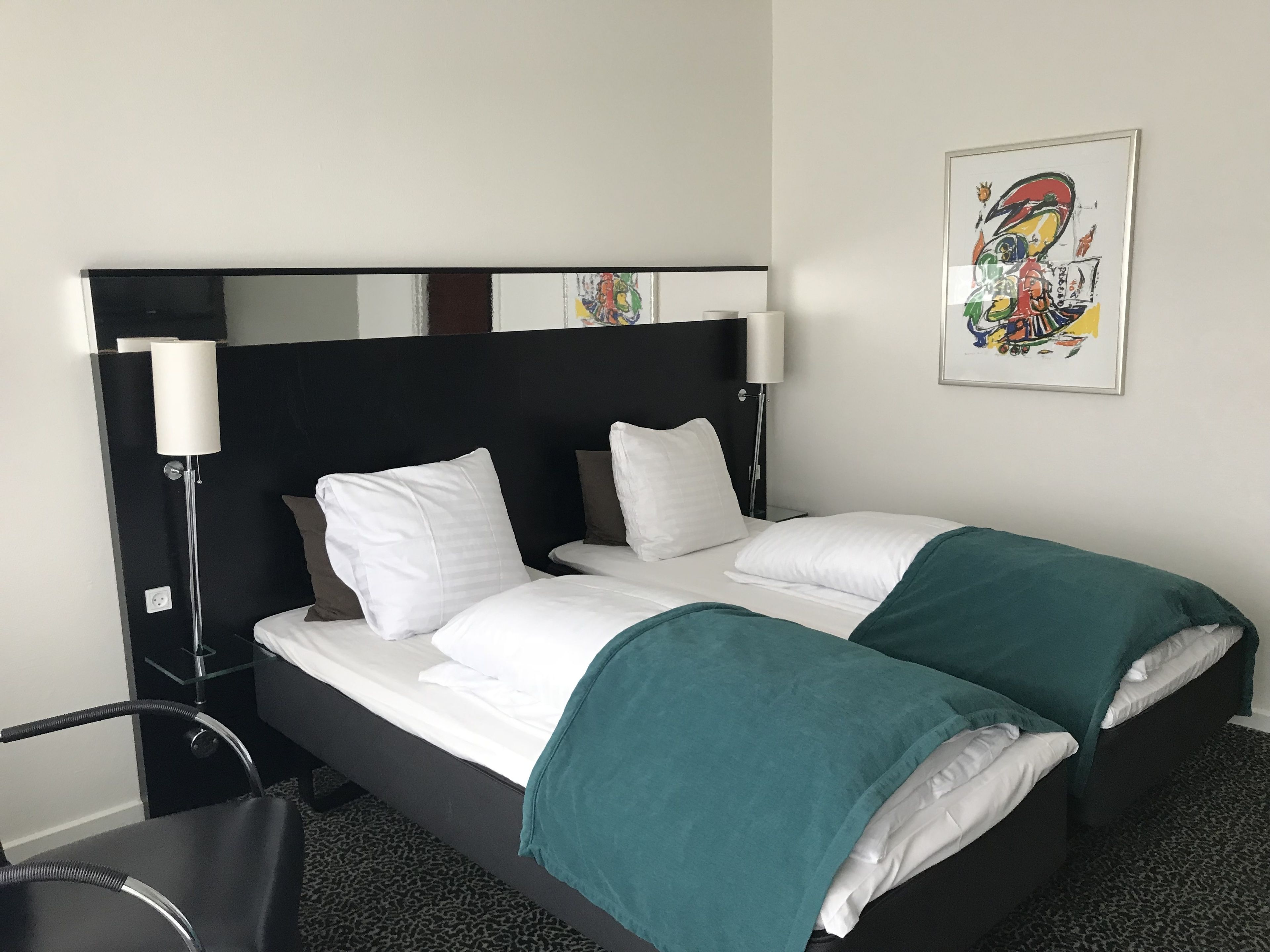 Et hotelværelse med sort sengegavl, hvidt sengetøj, et turkis tæppe, et enkelt natbord med lampe, og et farverigt abstrakt maleri på væggen.