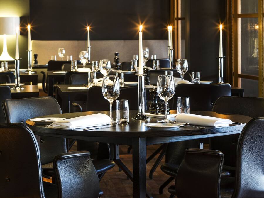 Un restaurante con sillas de cuero negro, mesas dispuestas con servilletas blancas, copas de vino y velas.