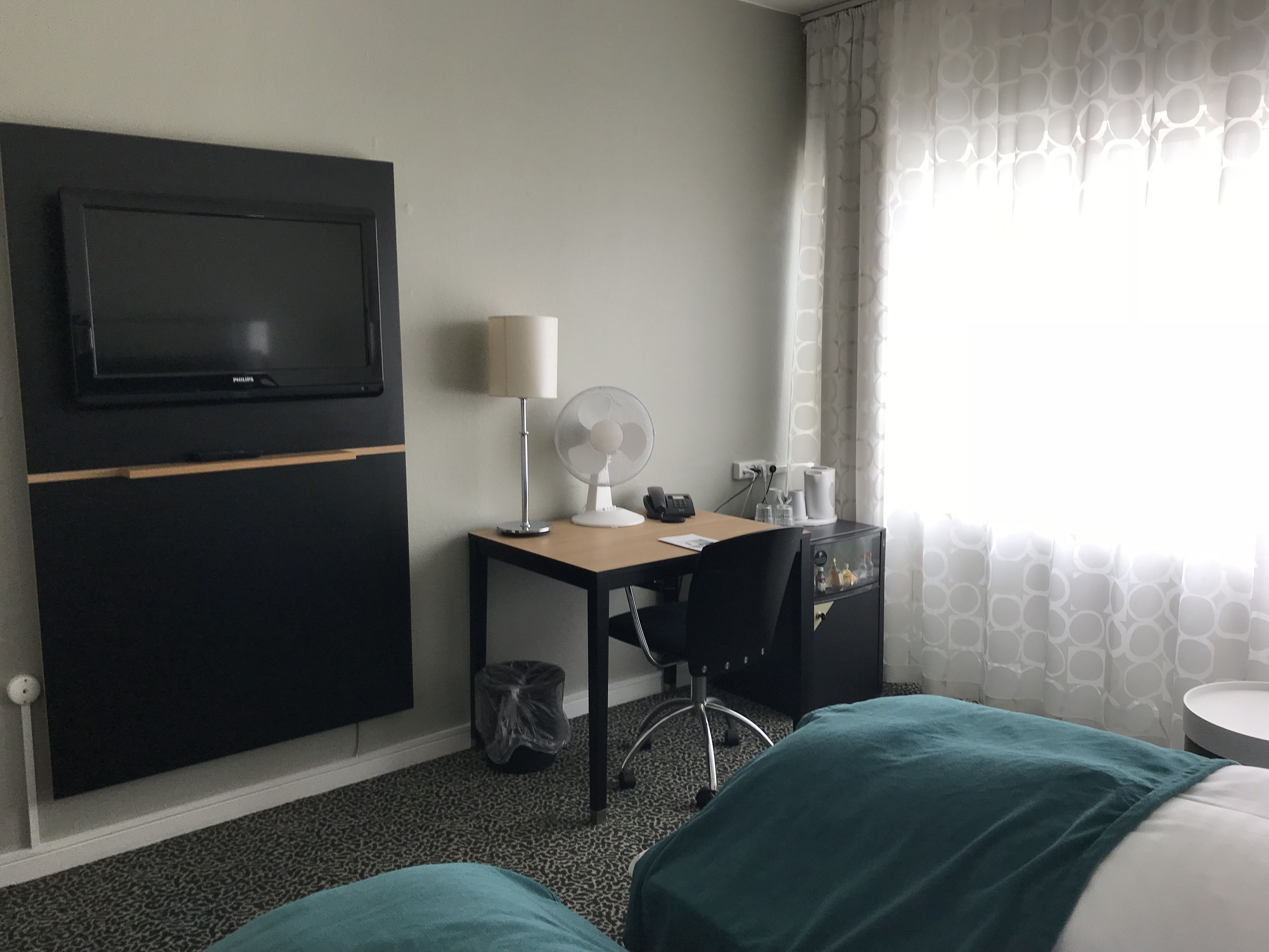 A hotel room with a bed, a desk with a lamp, a fan, a TV, and a window with curtains.