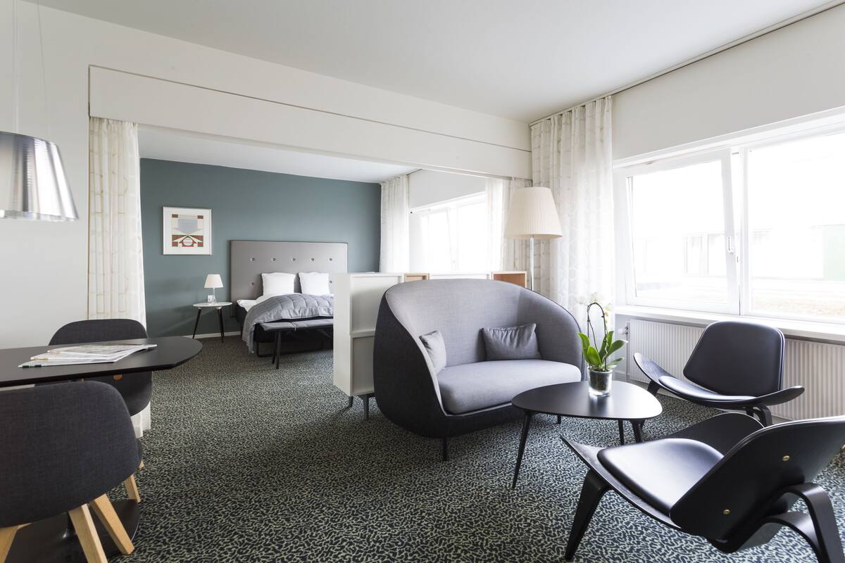 A hotel room with a bed, a grey sofa, a dining table with chairs, and a small table with a lamp and plant.