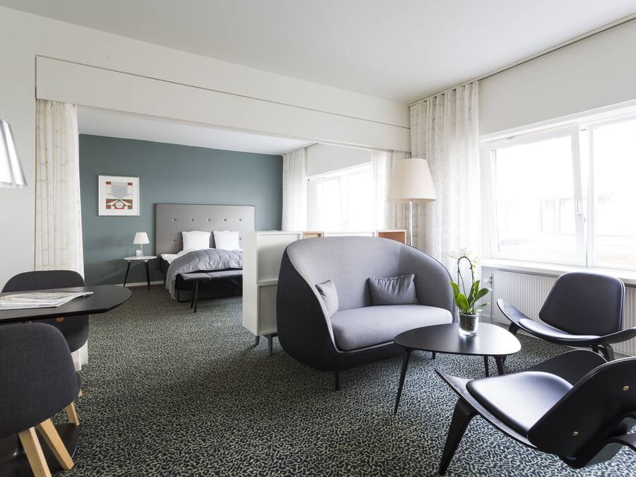 Et hotelværelse med en seng, en grå sofa, et spisebord med stole og et lille bord med lampe og plante.