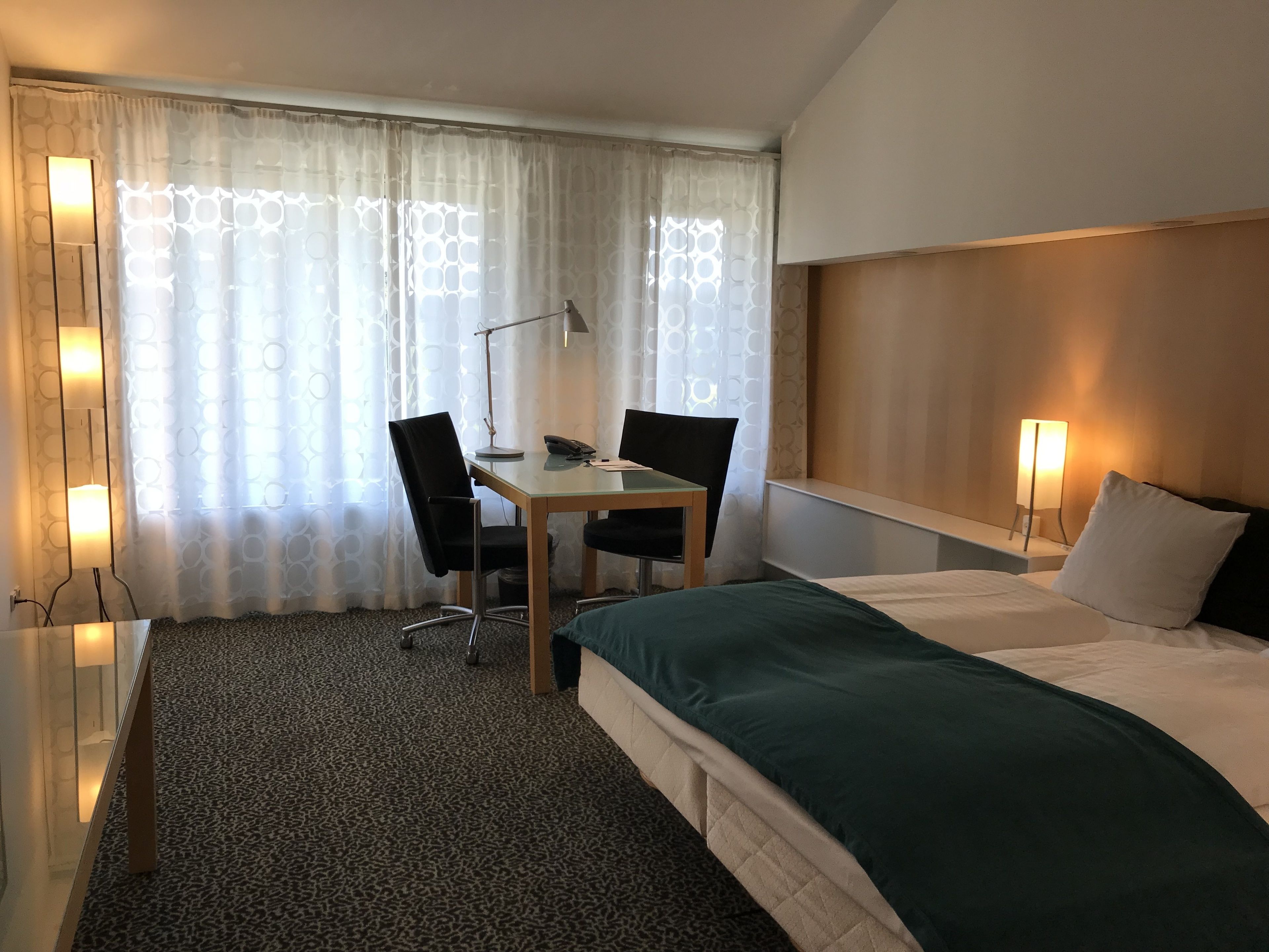 Una habitación de hotel con cama, escritorio, dos sillas, lámpara y mesita de noche.