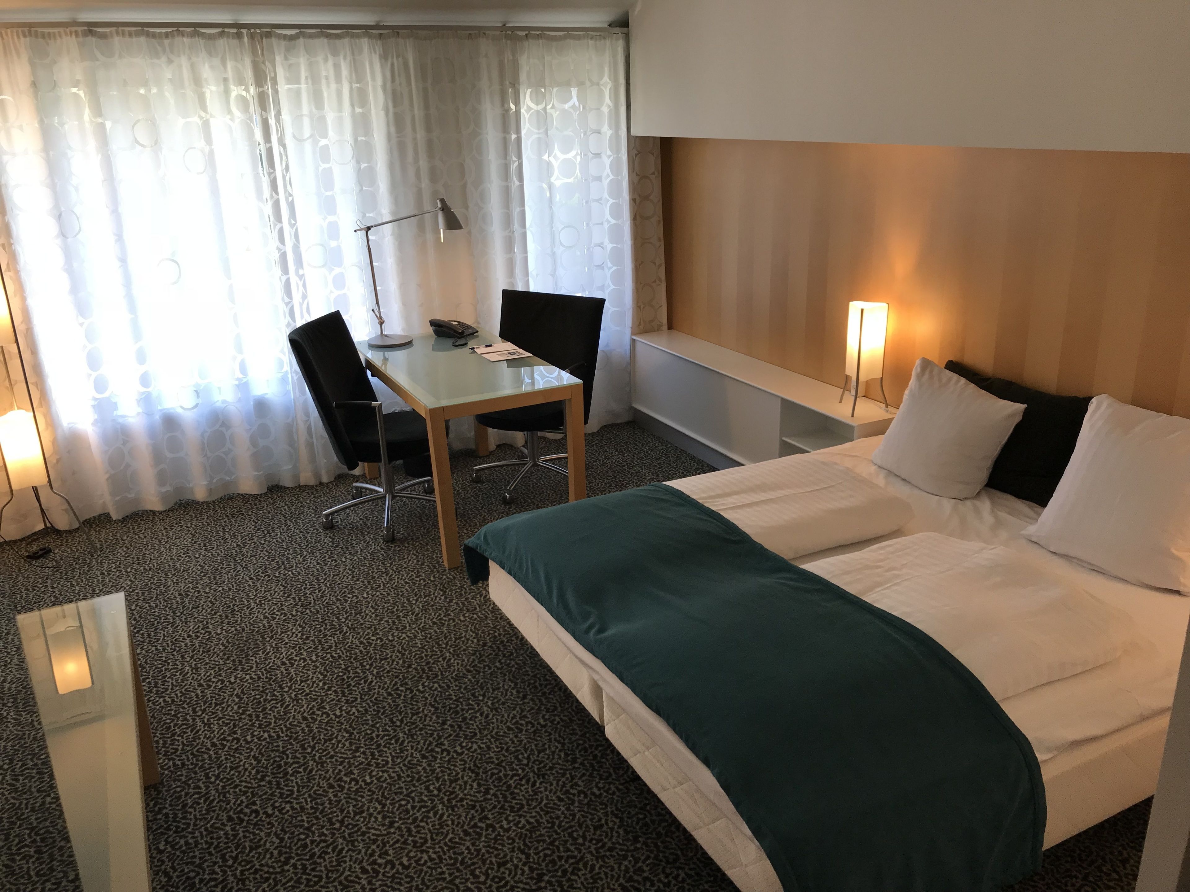 A hotel room with a bed, a desk, two chairs, a lamp, and a window with curtains.