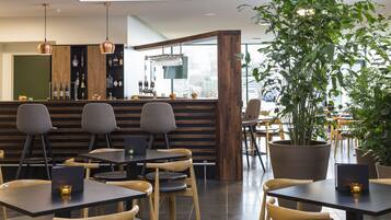 A modern restaurant with wooden tables and chairs, a bar area with bottles and glasses, and large potted plants.