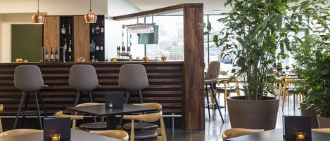 A modern restaurant with wooden tables and chairs, a bar area with bottles and glasses, and large potted plants.