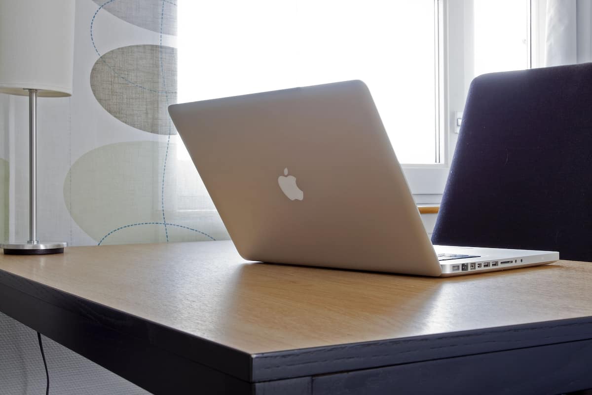 A laptop on a wooden desk near a window with a curtain.