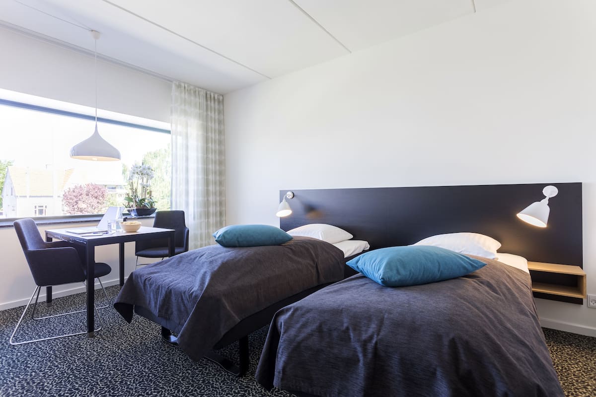 A hotel room with two beds, a desk, chairs, and a large window with curtains.
