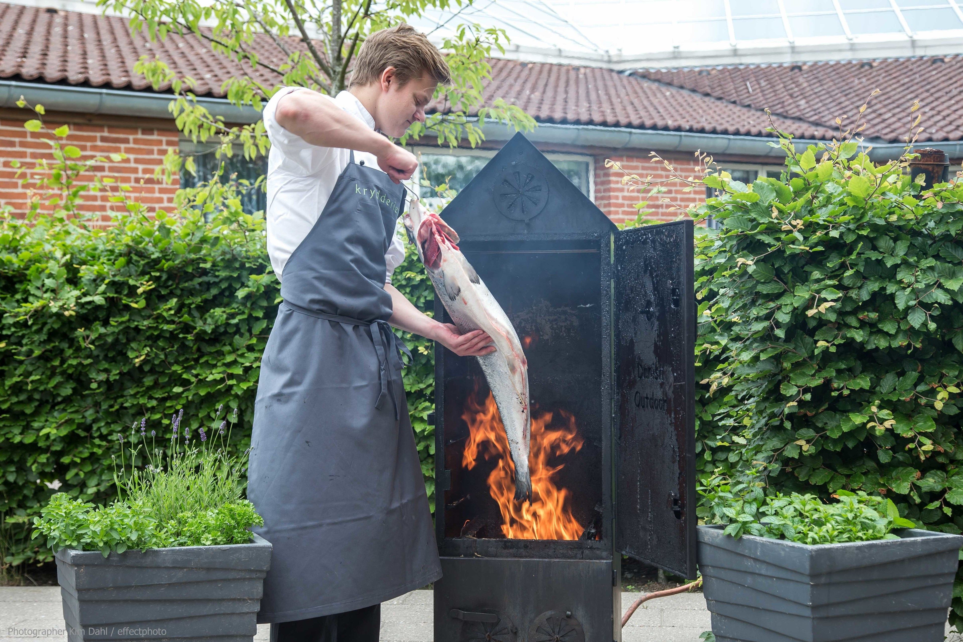 A person grilling a fish outdoors with a brick building and greenery in the background.