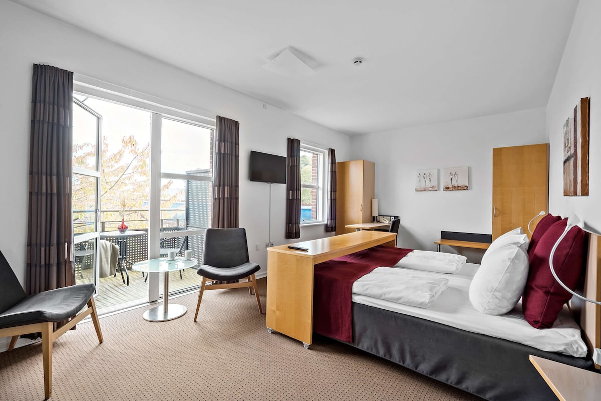A hotel room with a bed, a desk, a chair, a TV, and a balcony with a table and chairs.