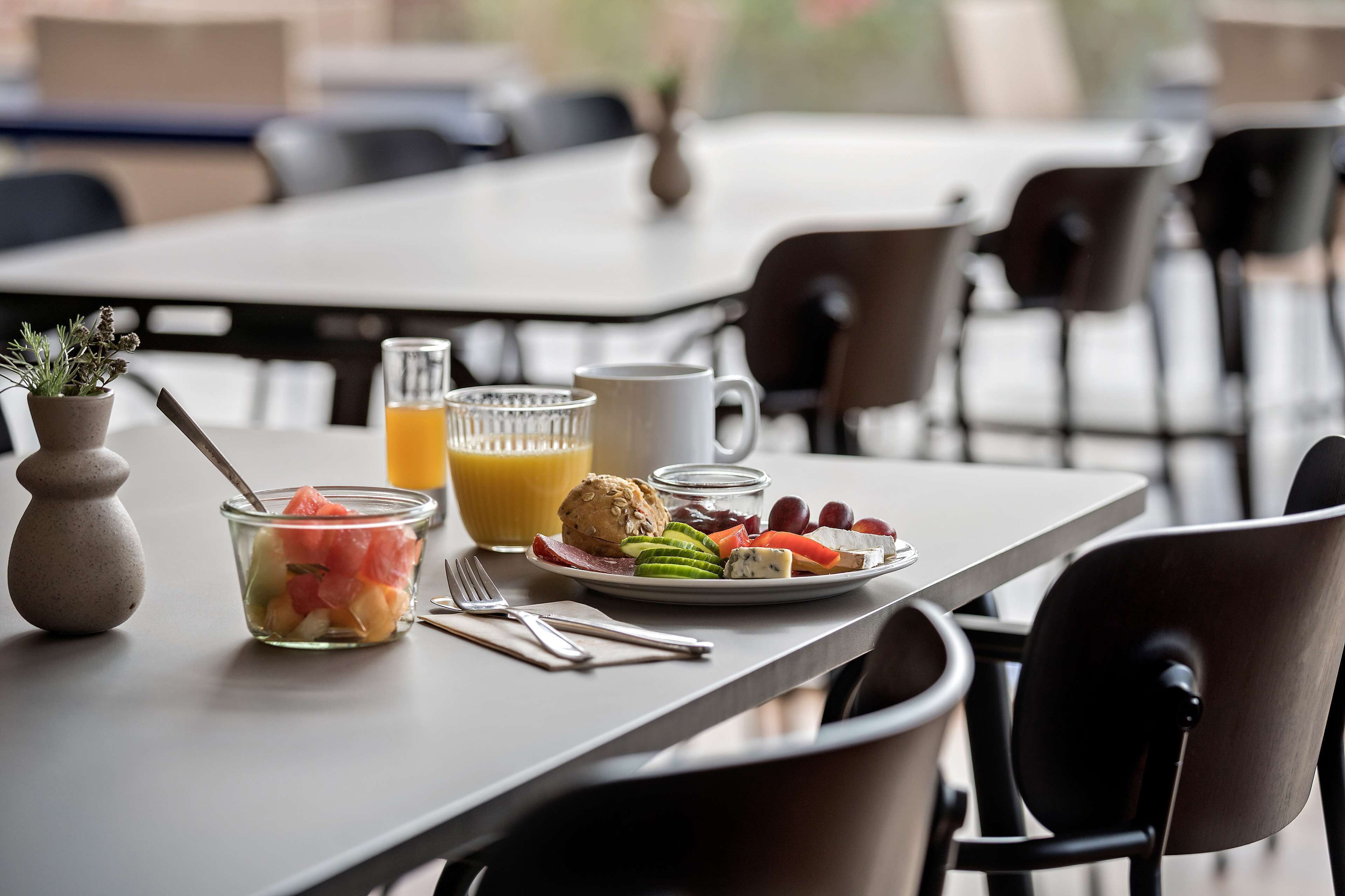 Una mesa de desayuno con un frutero, un vaso de zumo de naranja, una taza y un plato con diferentes alimentos.