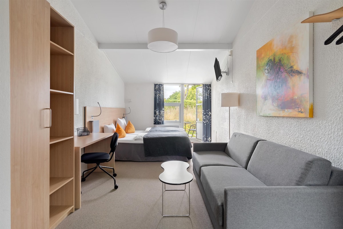 A modern bedroom with a bed, a desk, a grey sofa, and a large window.