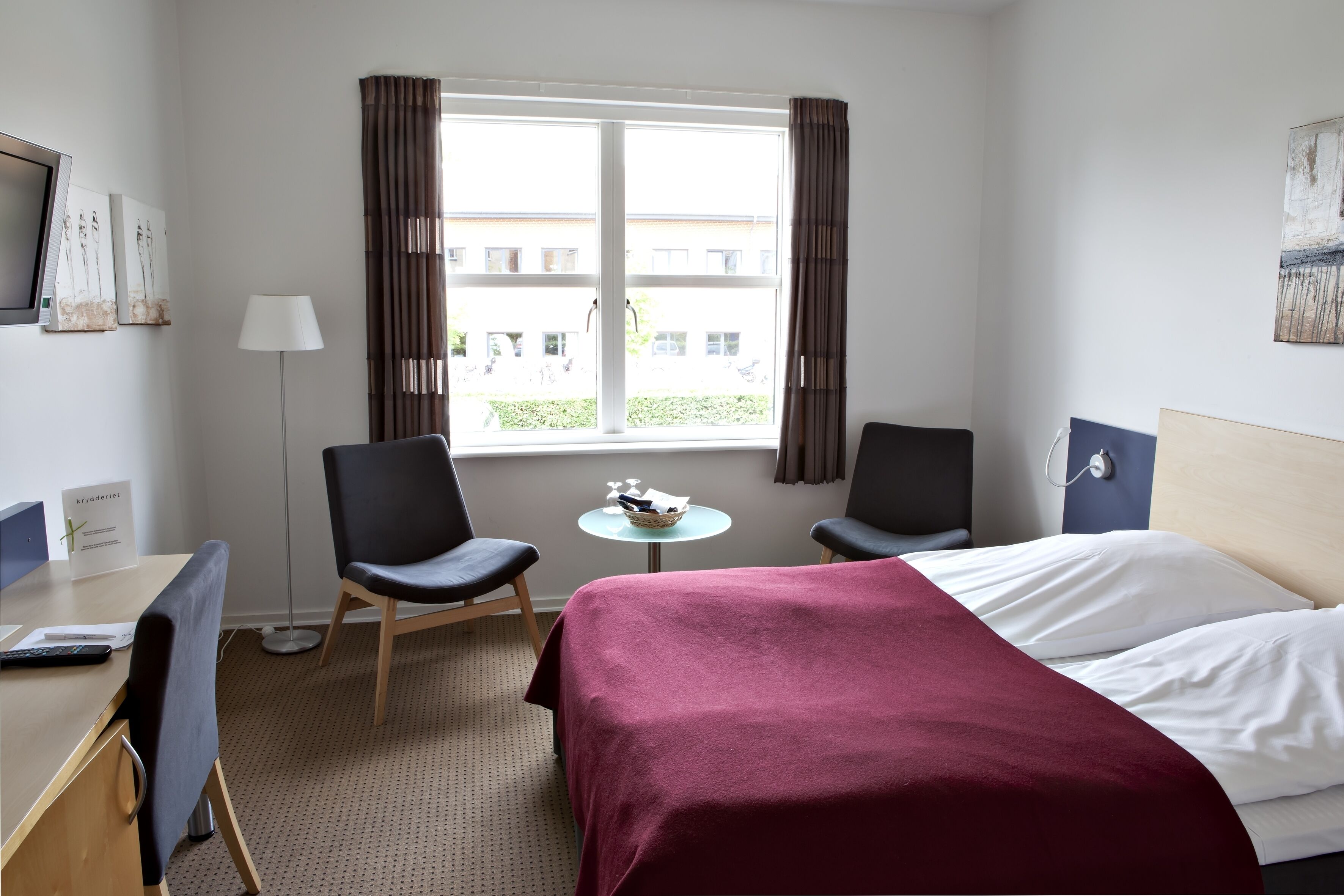A hotel room with a bed, two chairs, a desk, a lamp, and a window with curtains.