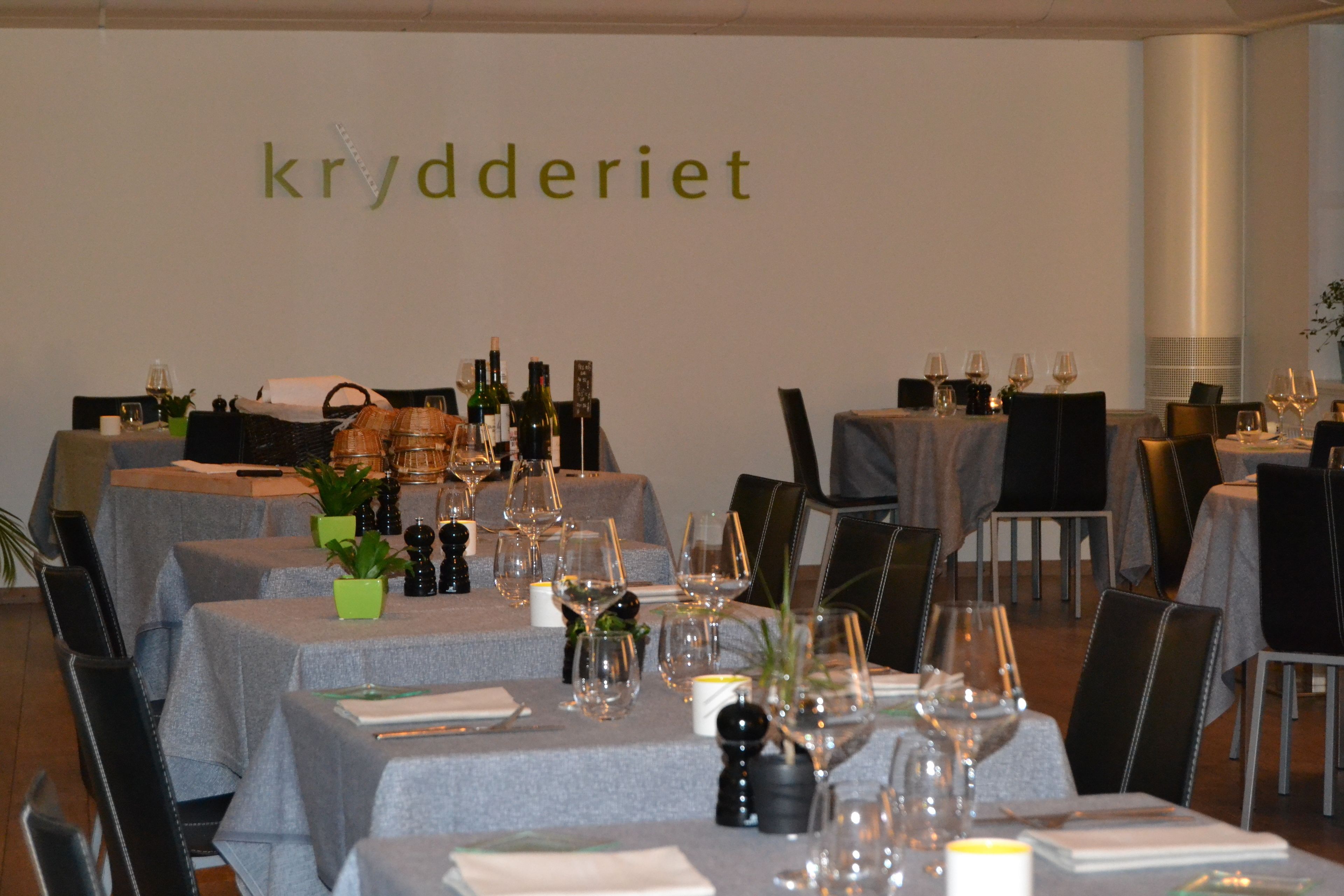 A restaurant interior with multiple tables set for dining, each with wine glasses, bottles, and small plants.