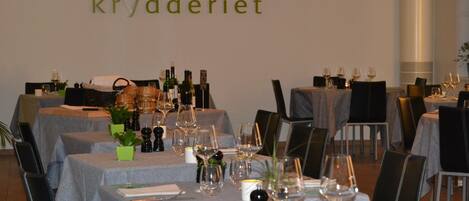 A restaurant interior with multiple tables set for dining, each with wine glasses, bottles, and small plants.