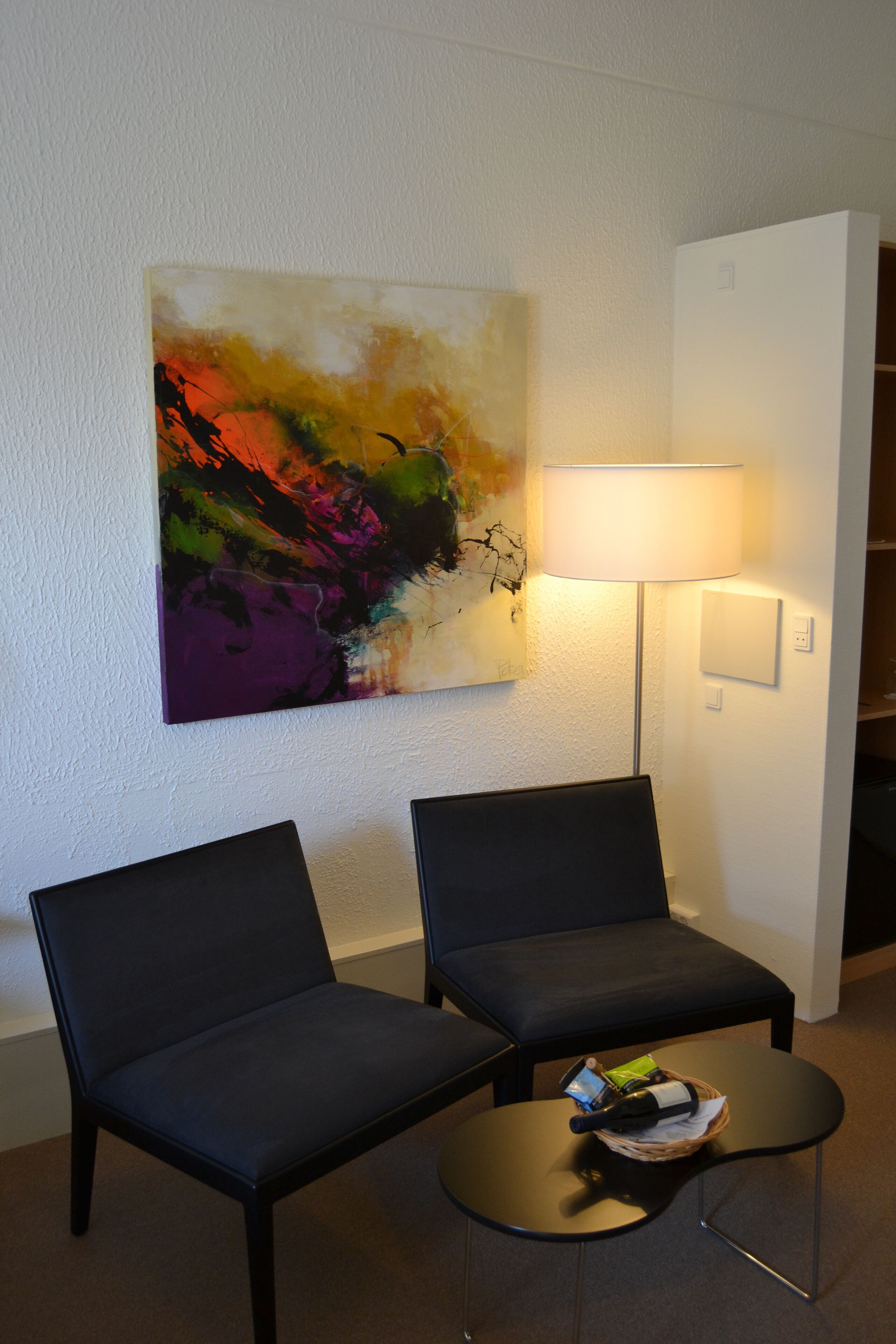 Una habitación con dos sillas negras, una mesita redonda con una cesta que contiene una botella y un libro, una lámpara de pie, y un cuadro abstracto y colorido en la pared.