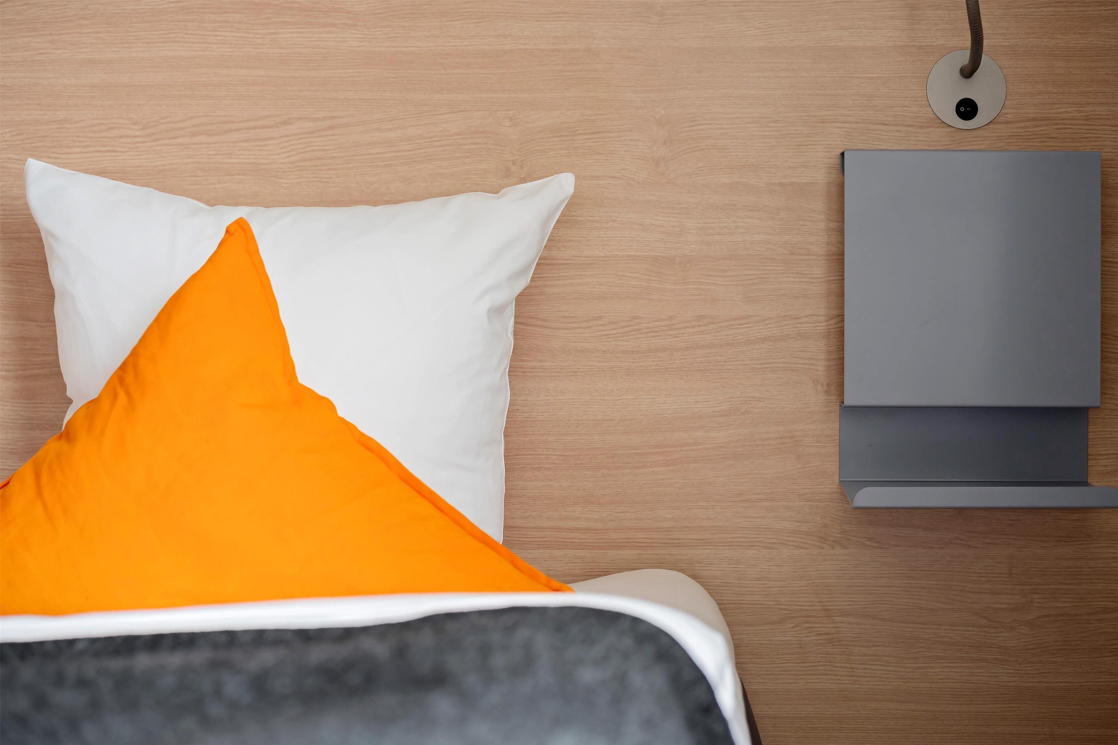 A neatly made bed with white and orange pillows, a grey blanket, and a grey bedside table with a lamp.