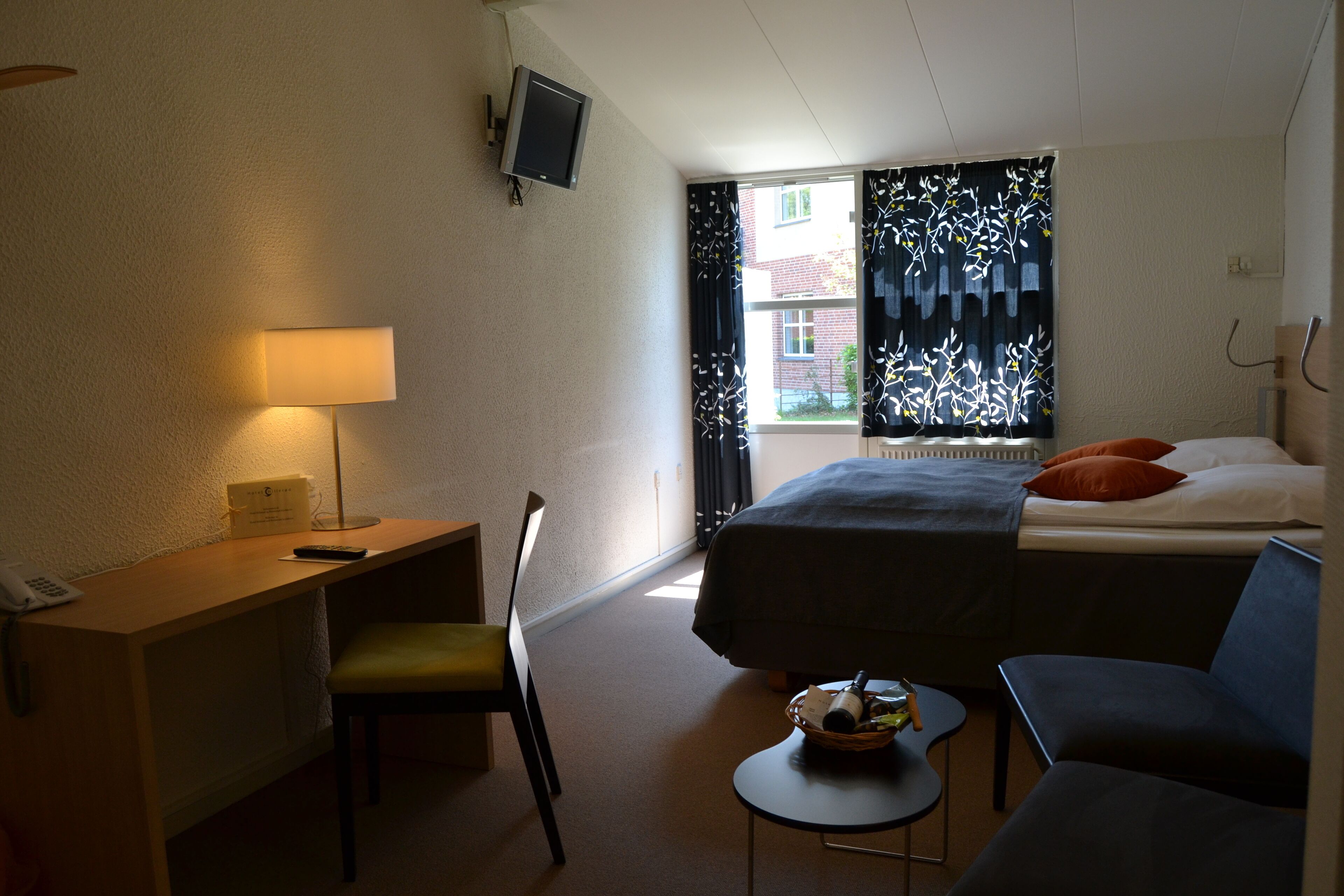 A hotel room with a bed, a desk, a chair, a small table, and a television.