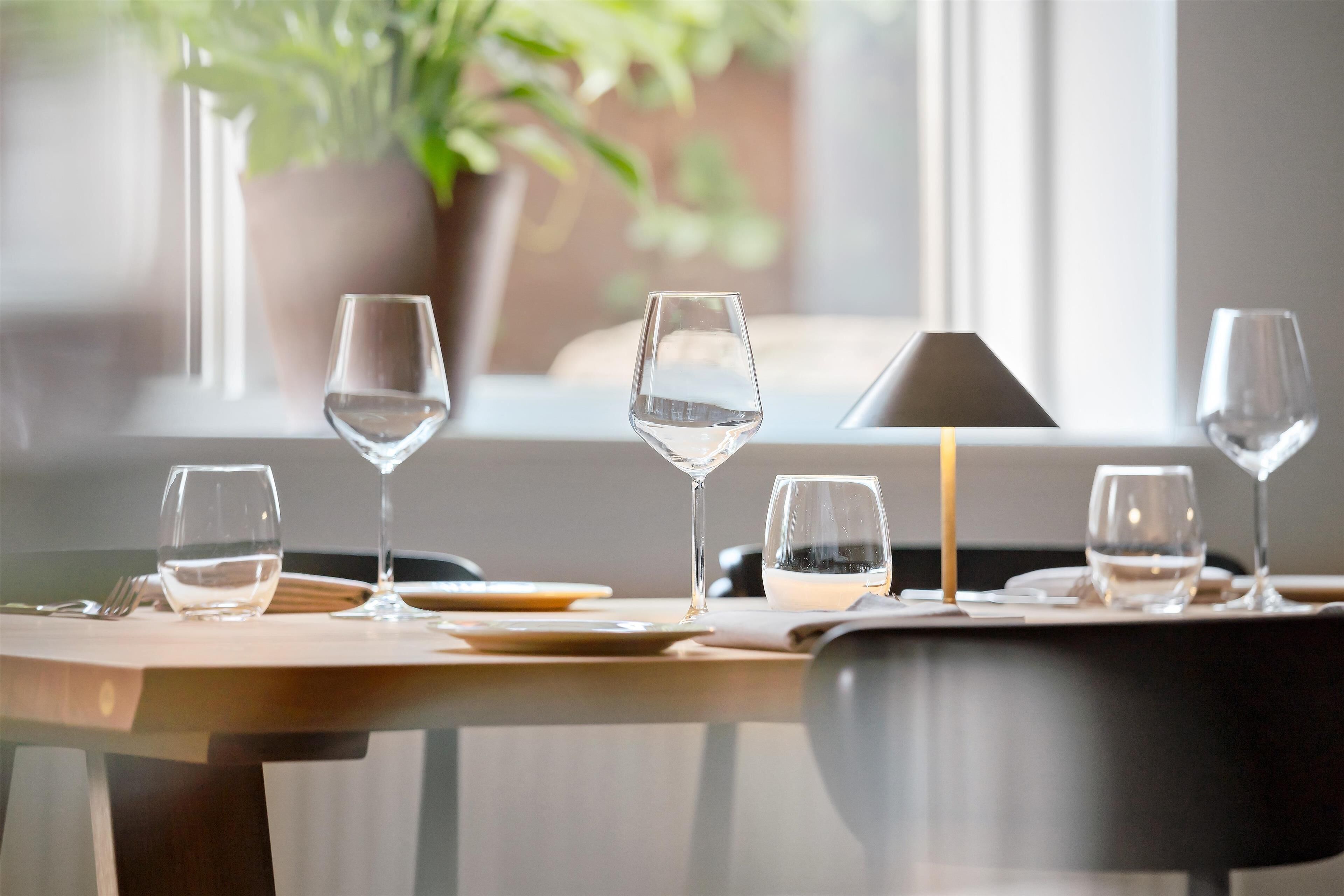 A dining table set with clear glasses and plates, a candle, and a potted plant in the background.