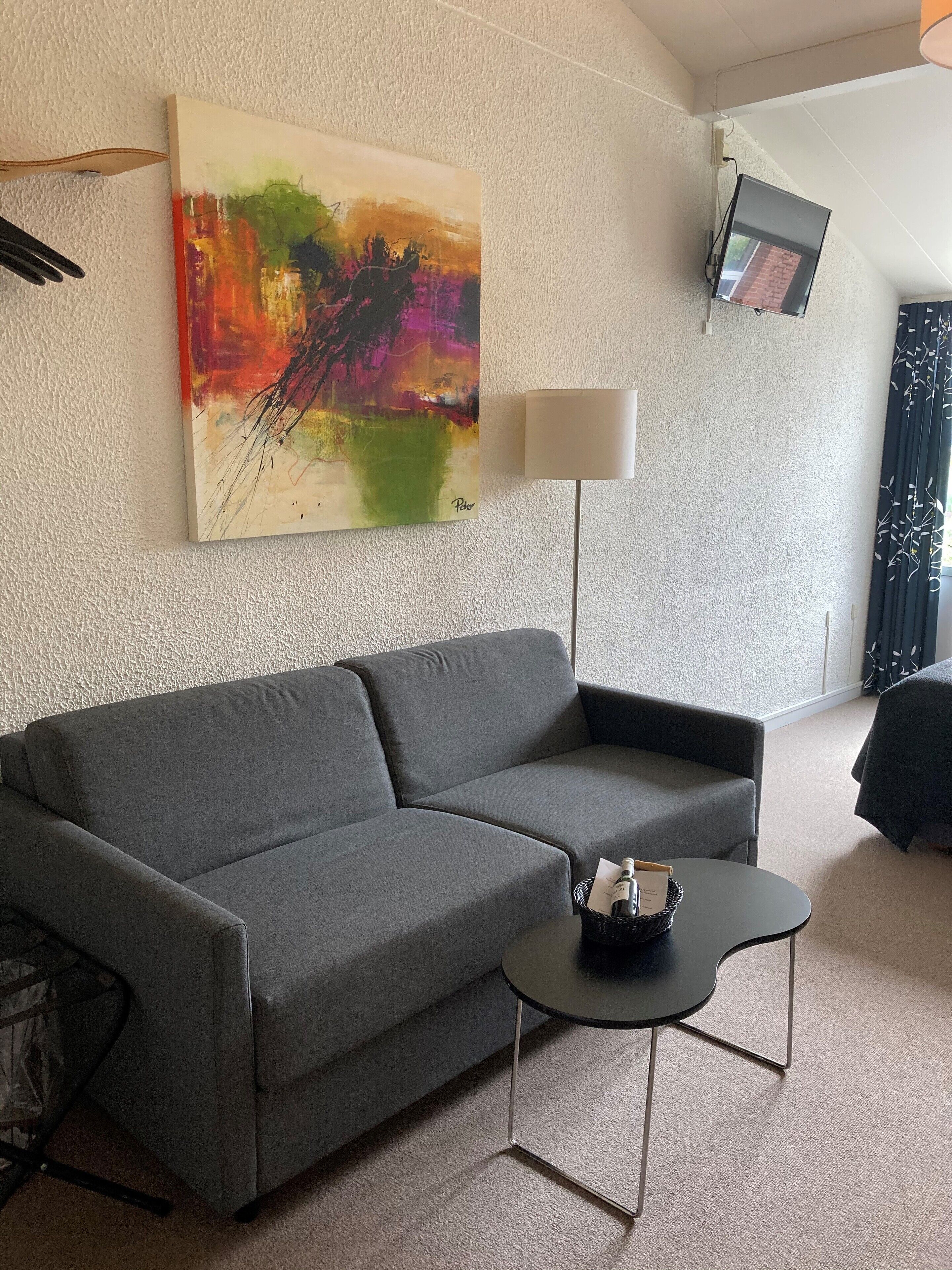 Una sala de estar moderna con un sofá gris, una mesita negra con una cesta, una lámpara de pie y un cuadro abstracto de colores en la pared.