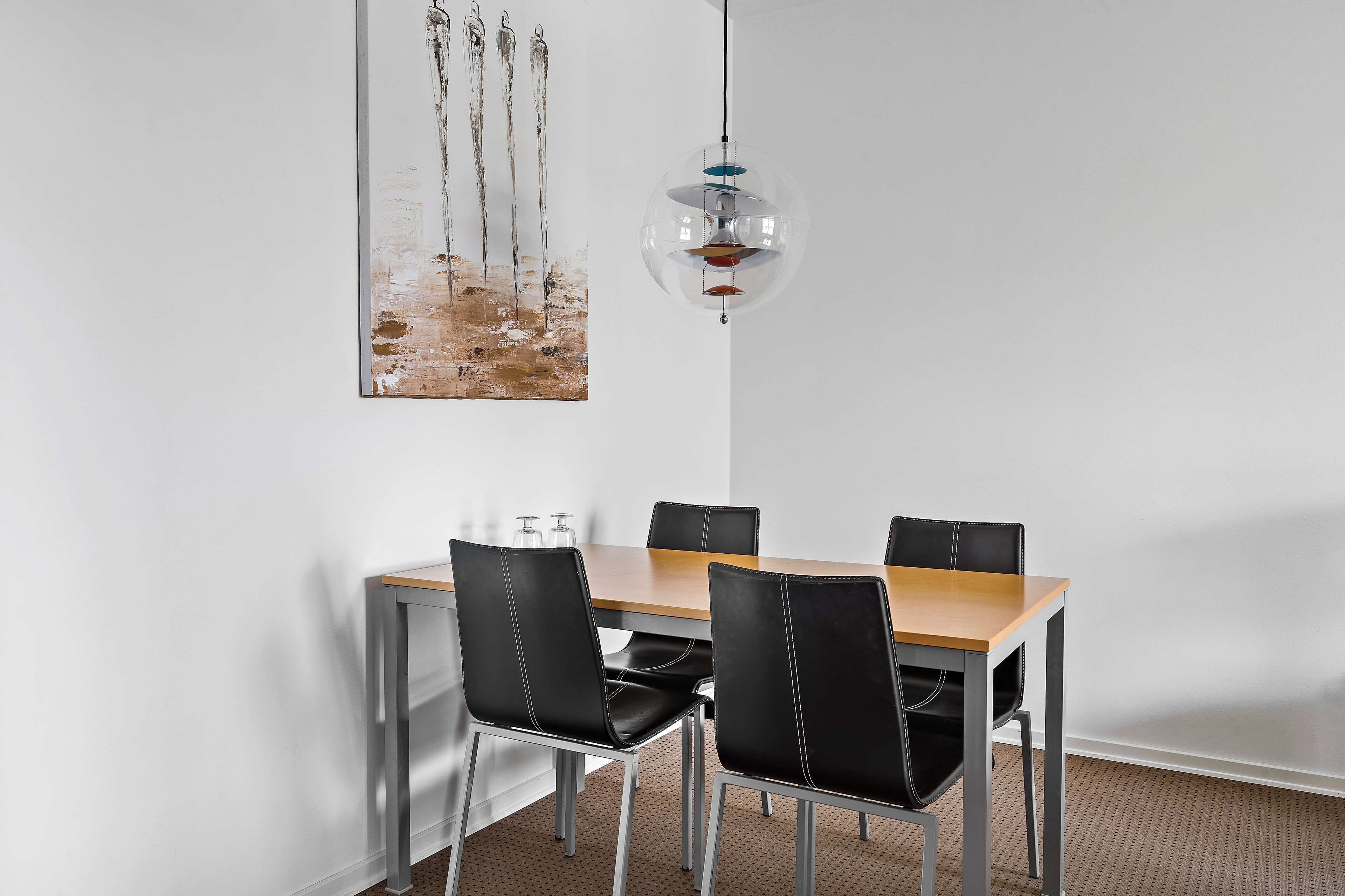 A dining area with a wooden table and four black chairs, a hanging pendant light, and a painting on the wall.