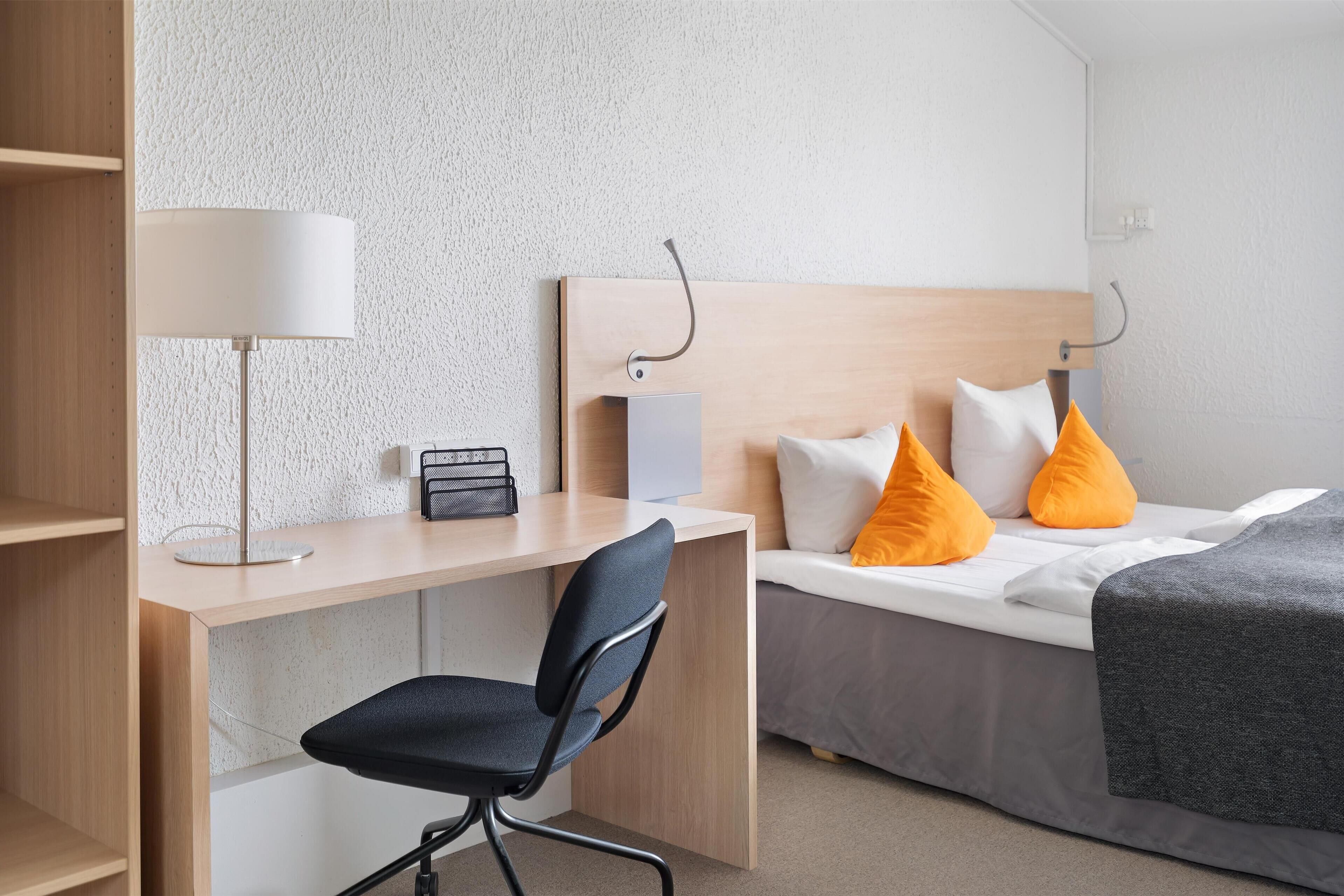 A hotel room with a wooden desk, a chair, a bed with orange pillows, a bedside table, and a lamp.