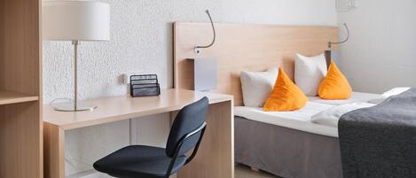 A hotel room with a wooden desk, a chair, a bed with orange pillows, a bedside table, and a lamp.